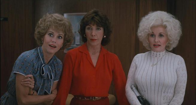 Jane Fonda, Lily Tomlinson and Dolly Parton look past the camera while stood in a 1980's office. Lily is wearing a red shift, Dolly is in a white jumper and Jane is wearing a blue and white tea dress. 