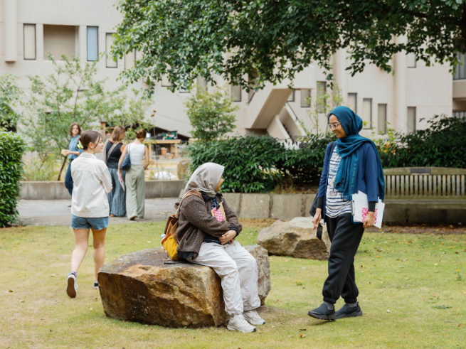 A grassed outdoor space with two large rocks in the foreground and a bench set further back. A woman is sitting on the nearest rock, looking up at another woman who is standing
