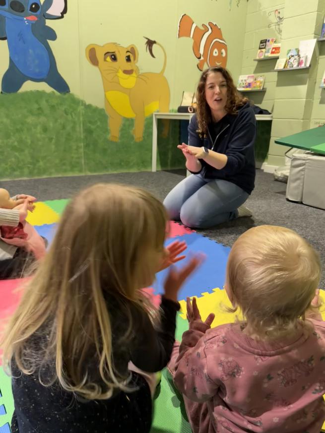 A female storyteller sat in front of children holding her hands in a book sign. The children's backs are to the camera and they are facing the storytelling mimicking her actions.