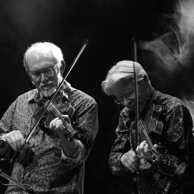 Black and white image. Bruce Molsky and Darol Anger play their violins on a low-lit, smoky stage.