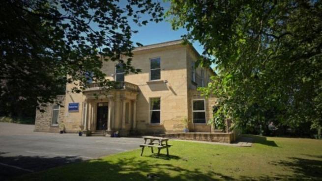 Large stone house with a columned entrance and tall sash windows, viewed from a lawn shaded by mature trees. A picnic bench stands on the grass in the foreground, with a driveway and entrance area to the left. The building is framed by leafy branches against a blue sky.