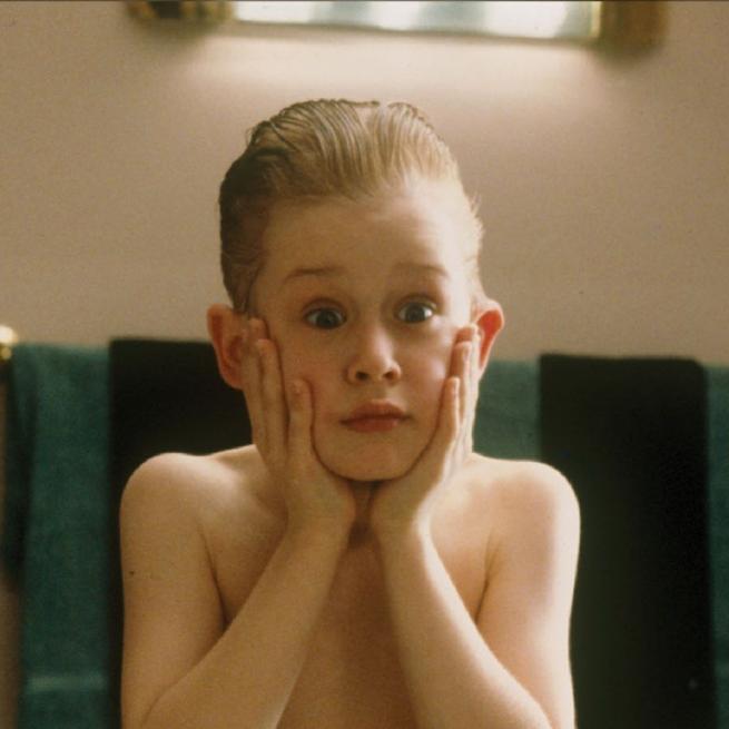Still from Home Alone, with a young boy in a bathroom with slicked back hair and two palms held to his face, pulling a shocked expression in the mirror with raised eyebrows.