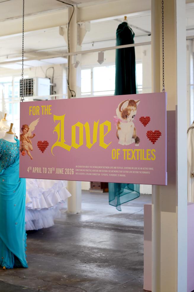 A lilac entrance board for the exhibition, featuring yellow text and graphics of kitsch kittens, hearts and cherubs. In the background is a selection of formal dresses on mannequins.