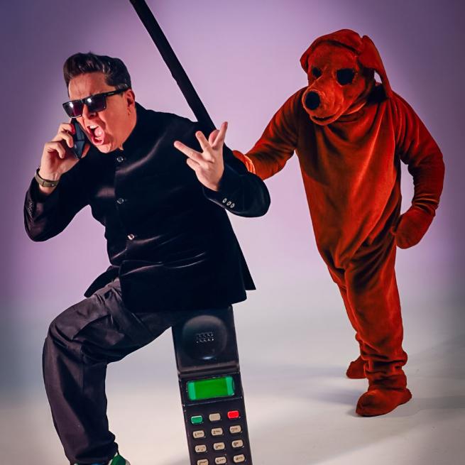 Dom Joly comically shouts into a mobile phone, wearing a shiny black jacket and sunglasses. A person in a reddish dog suit is behind him with a baton