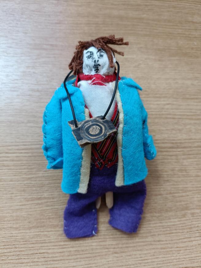 image shows a hand sewn peg doll with a light blue jacket and dark blue trousers. The face is drawn onto fabric with a black biro. The 'doll' has brown hair made from embroidery thread and has a camera around his neck.