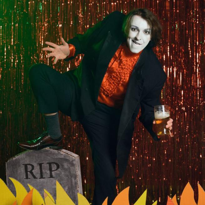 In white face paint and a frilly red shirt, Frankie Monroe grins and holds a pint of beer in front of a sparkly, red stage curtain, his foot rests on a prop gravestone amidst a row of cardboard flames.