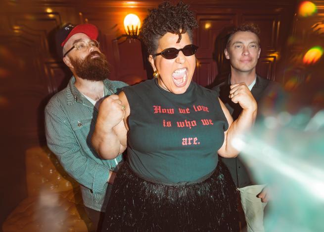 Three people in a warm, retro-lit room, with the central person wearing sunglasses, flexing and shouting, and a shirt that reads ‘How we love is who we are.