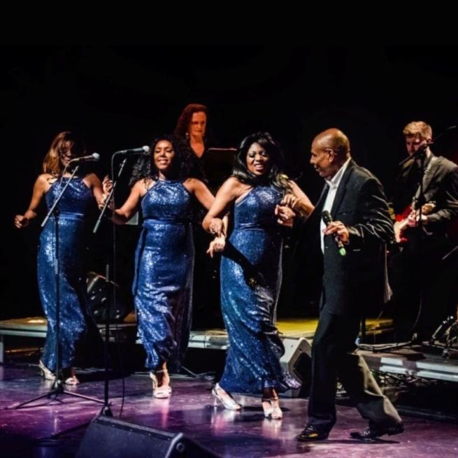 Three singers in sequined blue dresses dance by standing microphones with Roy Hemmings.