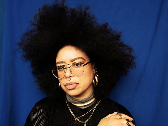 Person with voluminous curly hair and glasses stands against a blue background. They wear gold hoop earrings, layered necklaces, and a black top, conveying confidence.