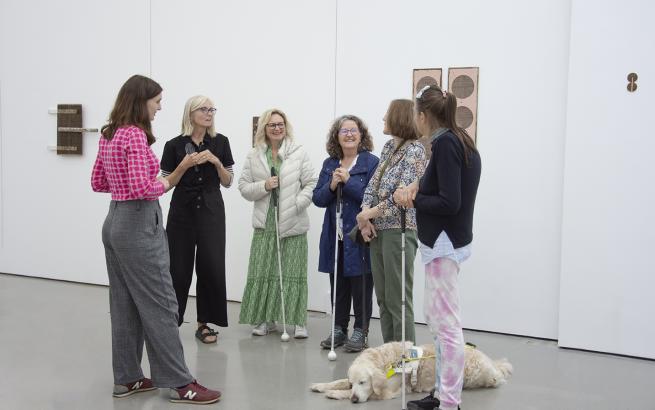 Six people standing in a gallery. they are talking and smiling. Three have white canes, one is holding a magnifying glass, and another has a guide dog resting at her feet.