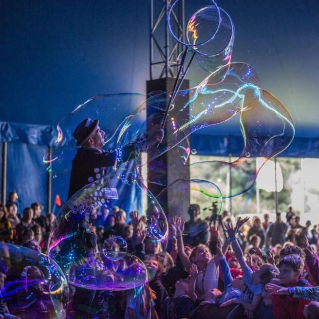 A man in a pork pie hat and polka dot waistcoat creates large bubbles on-stage in a festival tent, as children and families reach out in the audience.