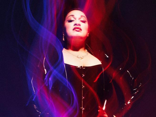 A woman posing in a dark setting, framed by vibrant, flowing streaks of red and blue light. She's wearing a dark corset and jacket, looking upward with a confident expression.