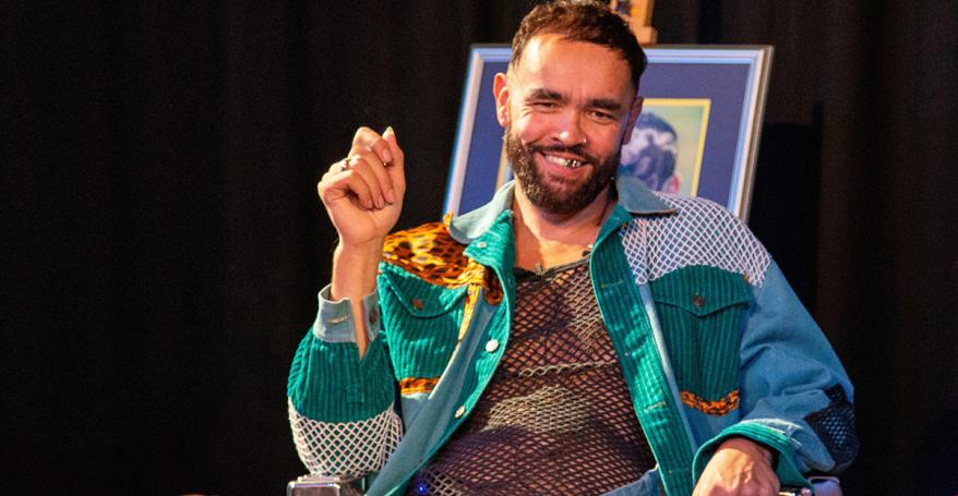  A man with a beard smiles while seated. He wears a colorful outfit and sits next to a stack of fabric. There's a framed picture and a fish decoration in the background.