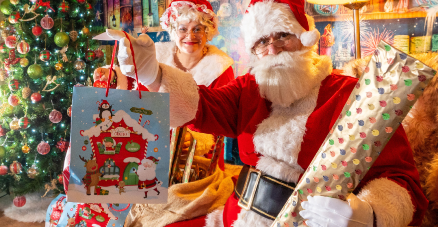 Santa and Mrs Claus sat in a wood cabin next to a decorated Christmas tree. Santa holding two presents and smiling.