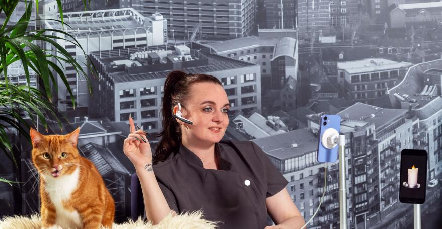 A receptionist wearing a spa uniform and headset; an orange cat is on the desk; a mobile phone with a candle emoji is also on the desk; a black and white backdrop of Leeds is behind the receptionist