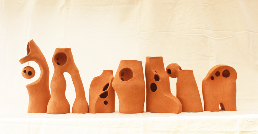 A row of abstract terracotta sculptures with organic shapes, rounded edges, and holes, displayed against a neutral fabric backdrop.