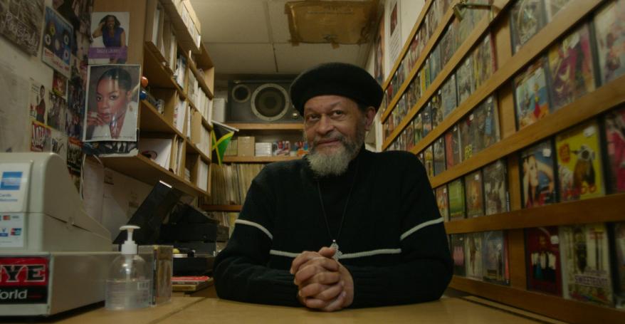 black record store owner owner wearing all black sat in his store surrounded by records