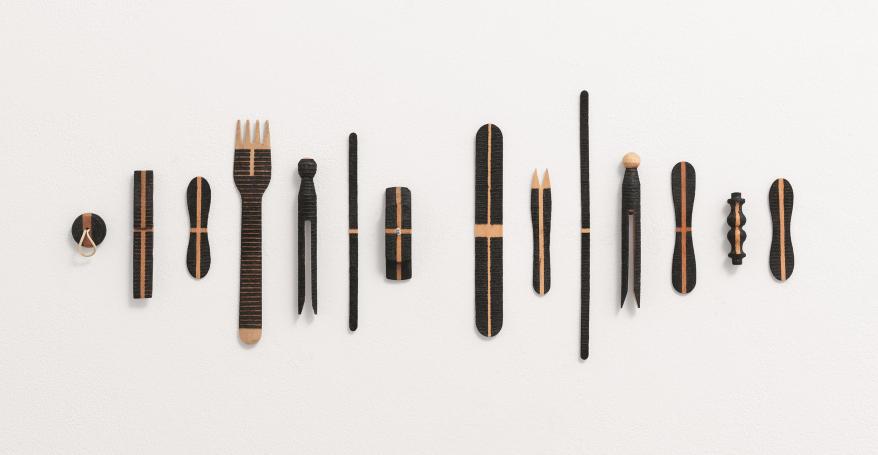 Fourteen everyday wooden objects, including a plug, chip fork, lolly stick, peg are arranged in a line on a wall. They have all been 'decorated' with different patterns of black lines, burnt by the sun.