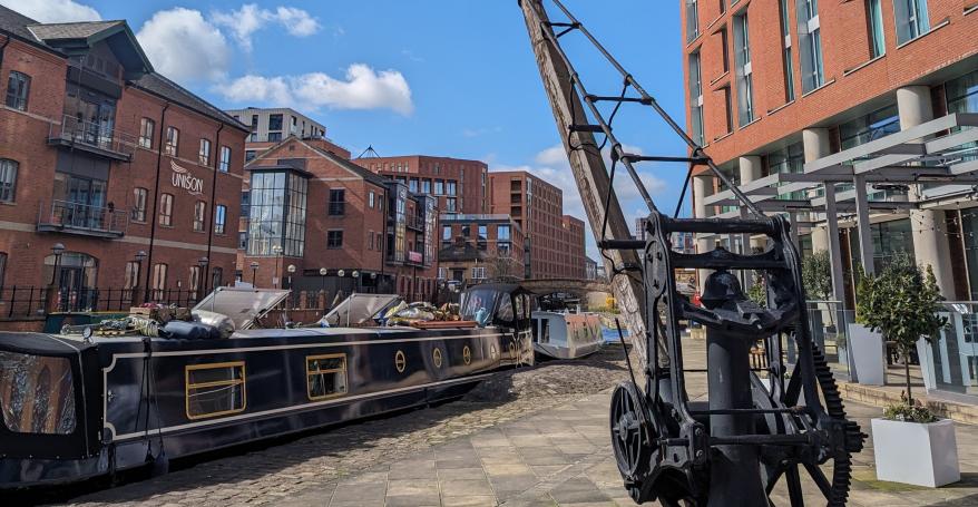A docked narrowboat next to a heritage crane and surrounded by buildings and blue sky at Granary Wharf in Leeds
