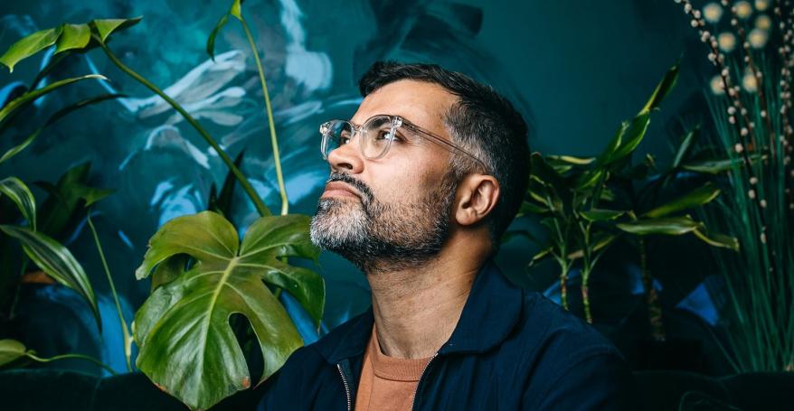 A man wearing glasses and a dark shirt stands in thought in front of a background of plants and vegetation.