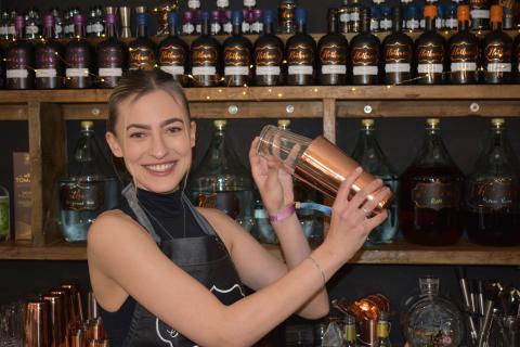 Photograph of Hotham's bar staff shaking a cocktail shaker. Behind her are rows of our award-winning gins.