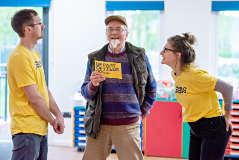 Three people stood together talking. In the middle a man is stood holding a yellow flier. On the left and right hand side two people are stood in yellow LEEDS 2023 t-shirts.