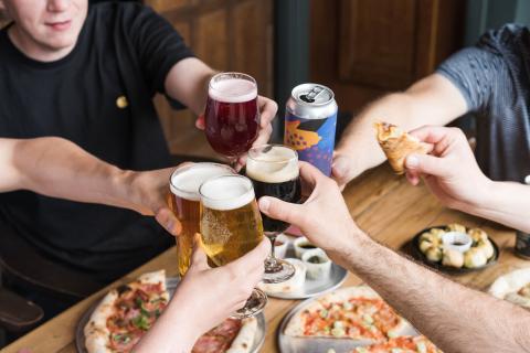 image shows people sitting at a table clinking beer glasses together. In the background the table is full of pizza.