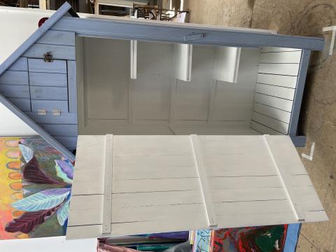 A blue tool shed, about the height and width of a tall, average build human. Painted white inside, with three wooden shelves. It has a pointed roof.