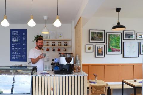 A barista stands in a cafe holding a cup of coffee. The cafe is a warm and friendly space with art on the walls.