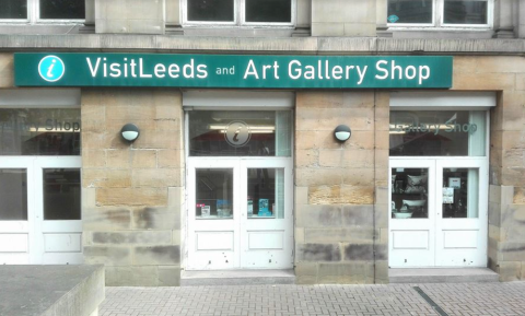 Green sign saying "Visit Leeds - Art Gallery Shop" above a brick building with three white doors.