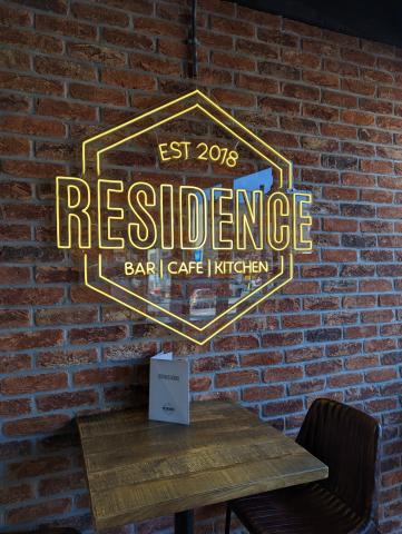 Brick wall. Hexagonal sign hanging up. Gold outline on sign and lettering. Sign reads: Residence Bar- Cafe- Kitchen Established 2018