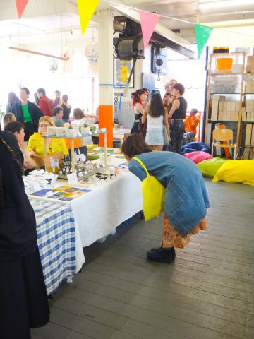 A room full of market stalls and bunting. In the forground a woman is bending down to look at a ceramics stall and in the background groups of people are standing and talking.