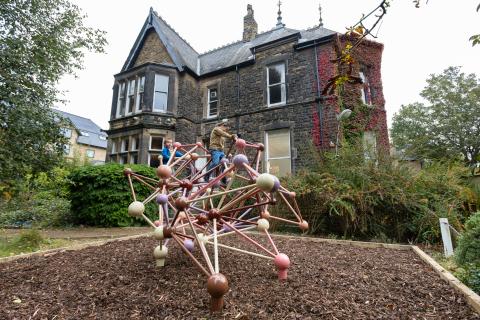 Two children playing on a colourful climbing frame with several unconventional geometric angles, in front of a Victorian manor