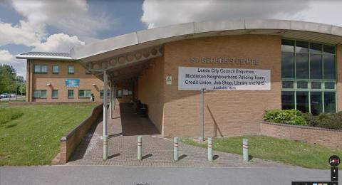 A building made of pale red-brown bricks with a curved grey roof above. To the left and in front of the building are green lawns. In front of the building are five pale green bollards with red stripes. There are silver letters on the side of the building which say 'St George's Centre'. A large white sign below with blue letters says 'leeds city council enquiries, middleton neighbourhood policing teams, credit union, job shop, library and NHS available here'.