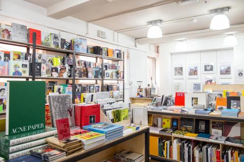 A well-lit bookshop features shelves filled with colourful books and magazines. The space is neatly organised with displays on tables and shelves, showcasing various art and photography publications. Bright overhead lights enhance the vibrant covers and titles on display. In the foreground, a large green book titled "ACNE PAPER" stands out alongside other eye-catching designs. The room has a clean, modern feel, with framed artwork on the walls, creating an inviting atmosphere for browsing.