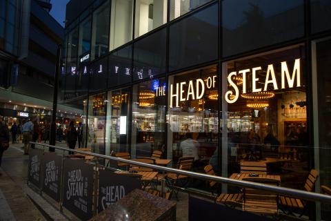 An exterior picture of the Head of Steam Bar on Park Row, Leeds. With large glass windows and the Name in illuminated letters on the outside. 