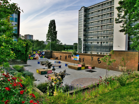 Wide landscape view of Roxby Community Garden and surrounding public square, showing colourful seating, murals, planting beds and nearby tower blocks in Lincoln Green.