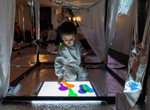 A child in a grey outfit interacts with a brightly lit color panel, touching colorful shapes in a dimly lit, reflective room filled with people.
