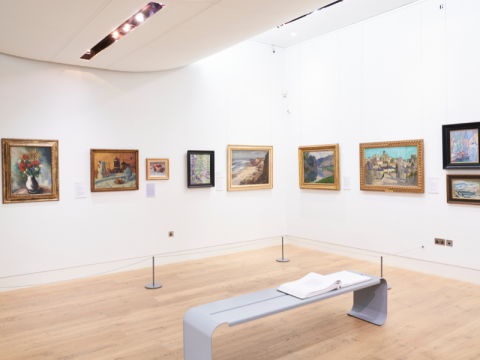 An art gallery room with 9 paintings hanging on white walls. There is a metal bench in the middle of the room