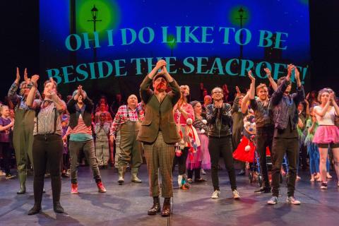 A large group of performers clapping in front of a backdrop that reads 'OH I DO LIKE TO BE BESIDE THE SEACROFT!'
