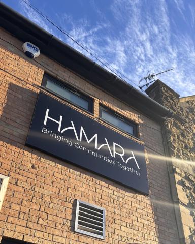 Brick building with a sign reading "HAMARA Bringing Communities Together" against a blue sky. Sunlight casts lens flares, creating a warm atmosphere.