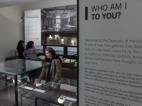 3 people stand in Treasures of the Brotherton museum. In the foreground there is an interpretation board that read 'Who am I to you?'  