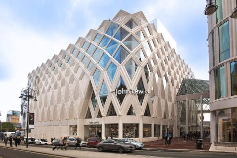 This image features the striking, contemporary architecture of a John Lewis department store building set against a bright sky. The facade is highly distinctive, covered in a geometric lattice pattern of beige diamond-shaped concrete or stone panels that frame large, angular glass windows. The street level shows busy pedestrian and vehicular traffic passing the ground-floor retail window displays, highlighting the store's urban location.