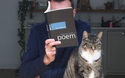 At a kitchen counter beside a tabby cat, Brian Bilston reads a book entitled 'how to write a poem', his face obscured behind its cover.