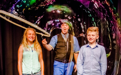 Two children stand either side of  a man with a large bubble wand, he is producing a large tube shaped bubble to cover them both. 