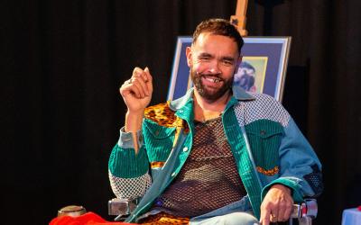  A man with a beard smiles while seated. He wears a colorful outfit and sits next to a stack of fabric. There's a framed picture and a fish decoration in the background.