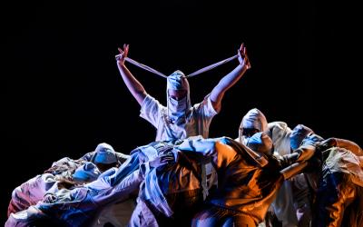 Dancer surrounded by a huddle wears a mask and looks directly at the camera