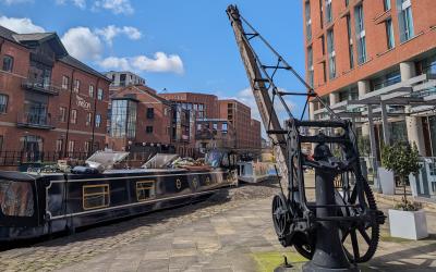 A docked narrowboat next to a heritage crane and surrounded by buildings and blue sky at Granary Wharf in Leeds