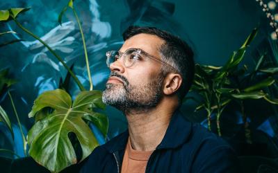 A man wearing glasses and a dark shirt stands in thought in front of a background of plants and vegetation.