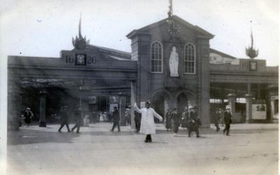 An image of Leeds City Square in 1926, with model moot hall erected for the tercentenary celebrations of Leeds getting its charter. A large building facade with a white statue in a central alcove (Queen Anne) stands in the centre. On either side the dates 1626 and 1926 are etched in what looks like stone. This is entrance to a station concourse. Pedestrians are milling about in period 1920s dress. A policeman in a white coat and gloves stands beneath the arch directing people.